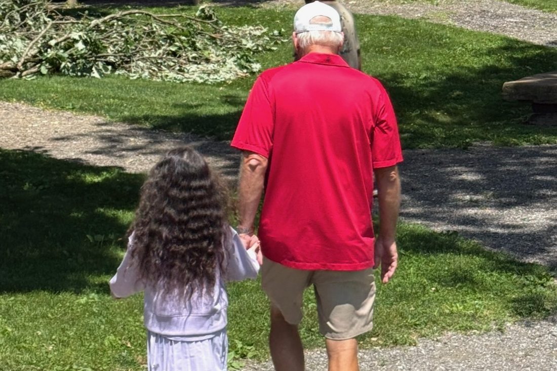 A grandfather and his granddaughter, seen from behind, walking hand in hand across a lawn